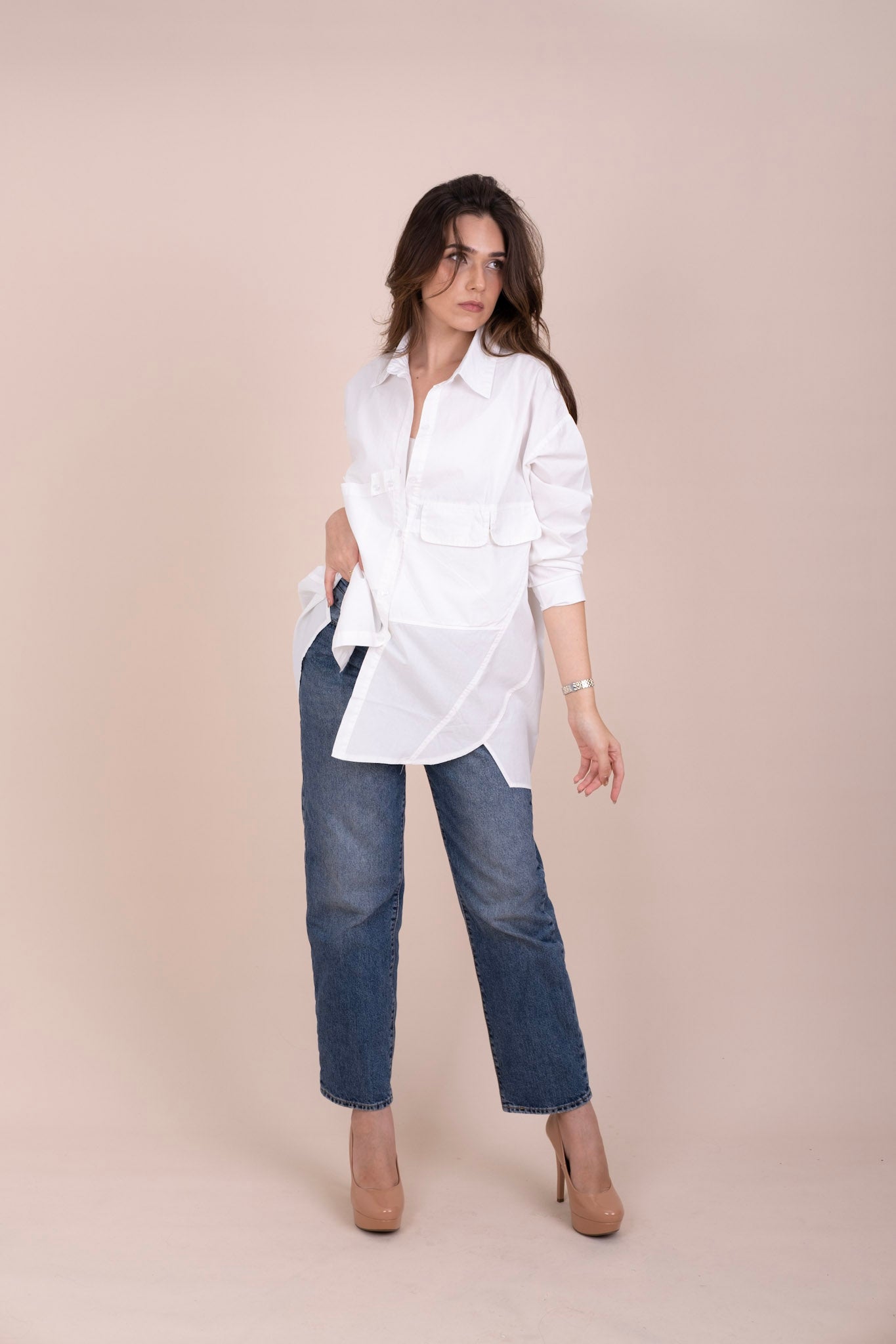 A woman modeling a white long-sleeve button-up oversized shirt with front flap pockets, paired with blue jeans and beige heels, against a solid light beige studio background.
