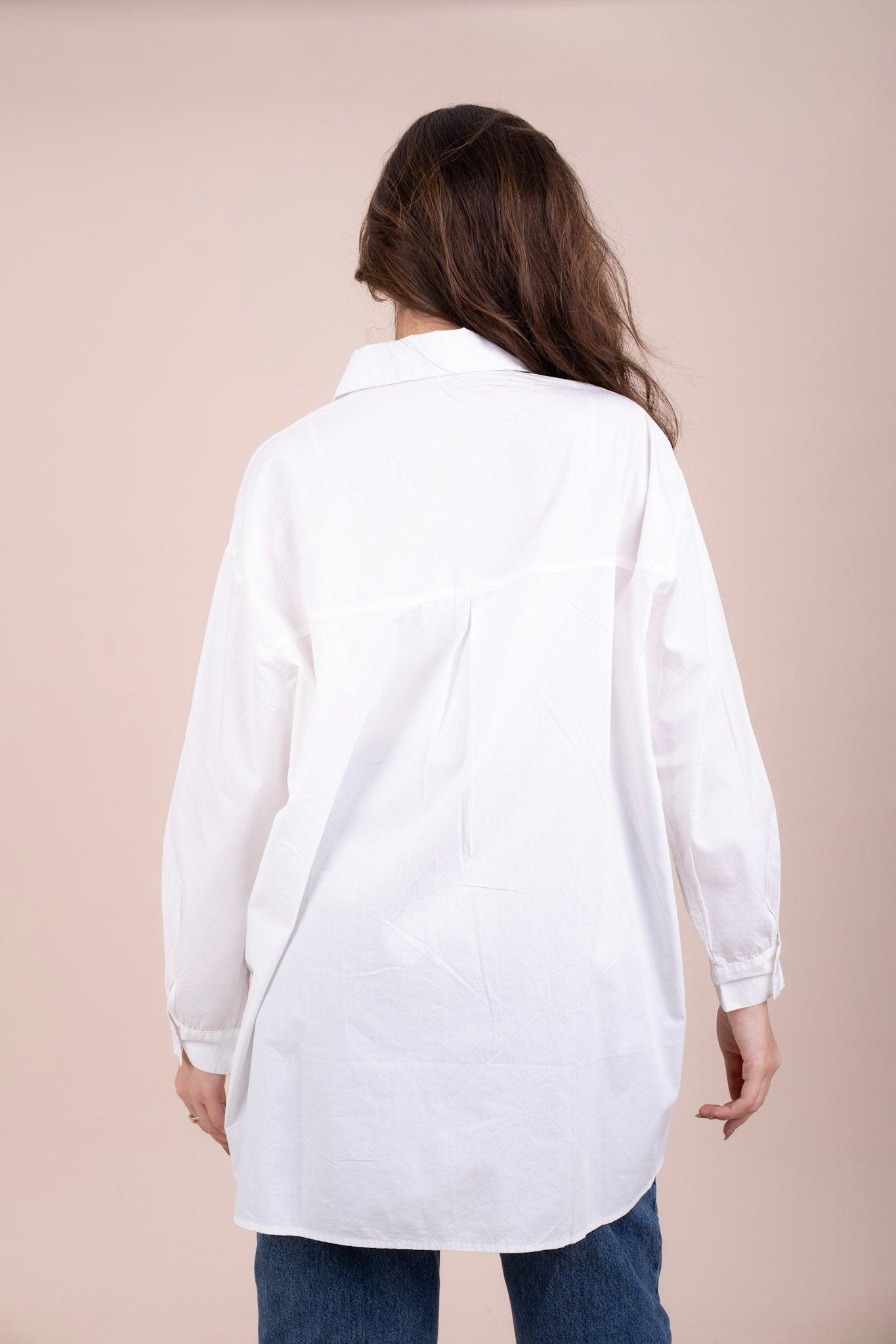 A woman viewed from behind wearing an oversized white long-sleeve button-up shirt and blue jeans, standing.
