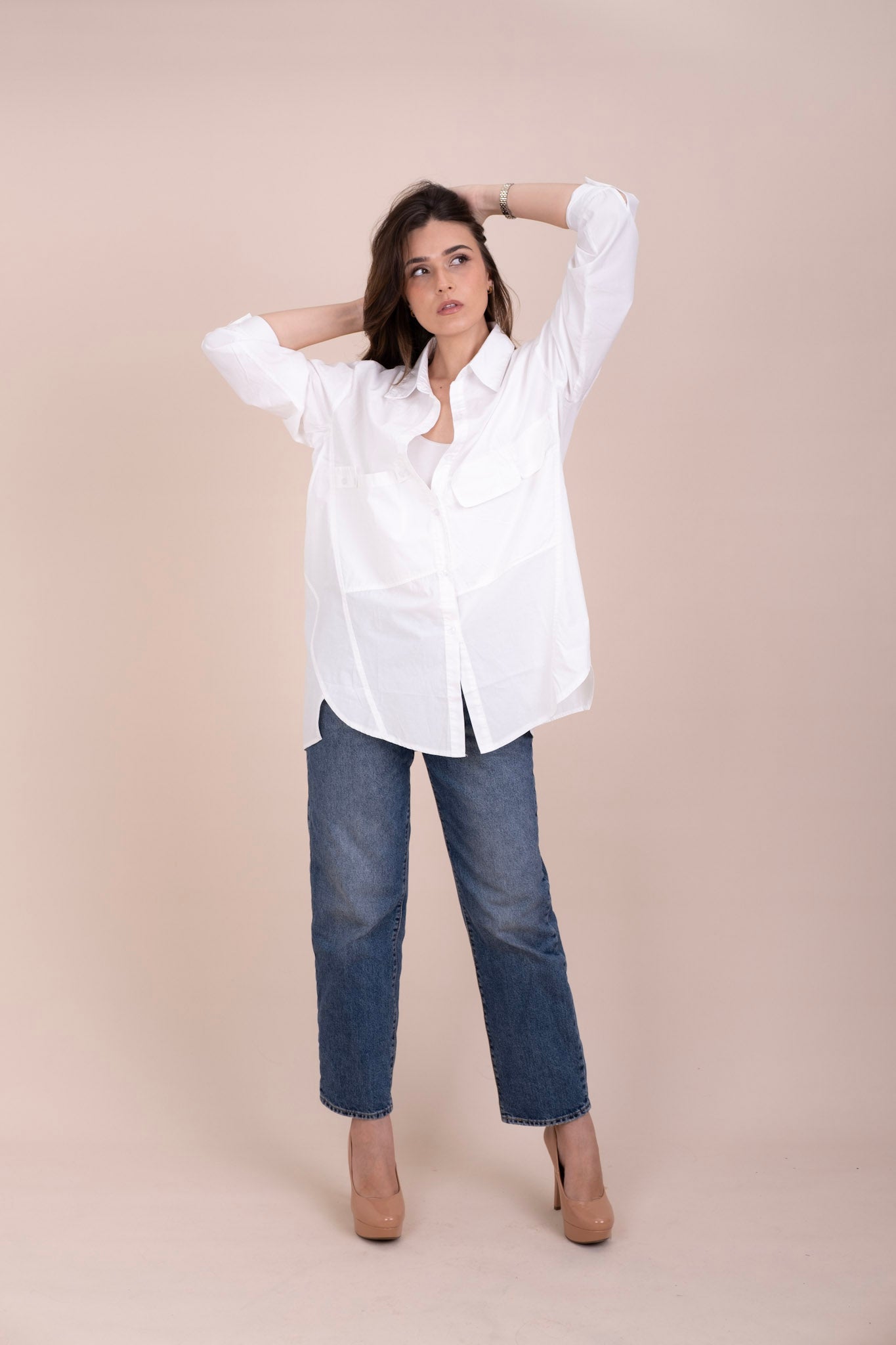 A woman modeling a white long-sleeve button-up oversized shirt with blue jeans and beige heels, posing against a solid beige studio backdrop.