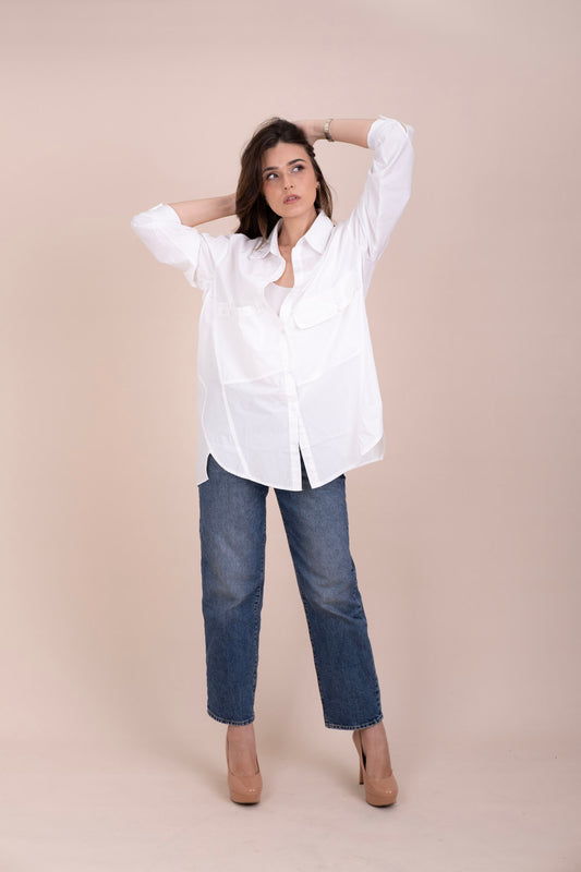 A woman modeling a white long-sleeve button-up oversized shirt with blue jeans and beige heels, posing against a solid beige studio backdrop.