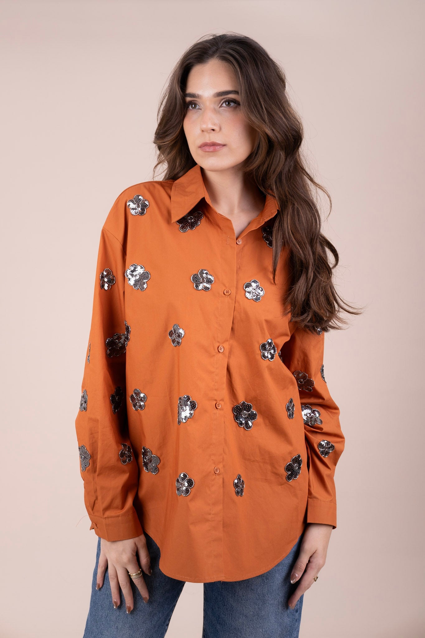 A woman in a long-sleeve button-up shirt in burnt orange, decorated with scattered silver sequined floral motifs, paired with blue jeans against a solid beige studio background.
