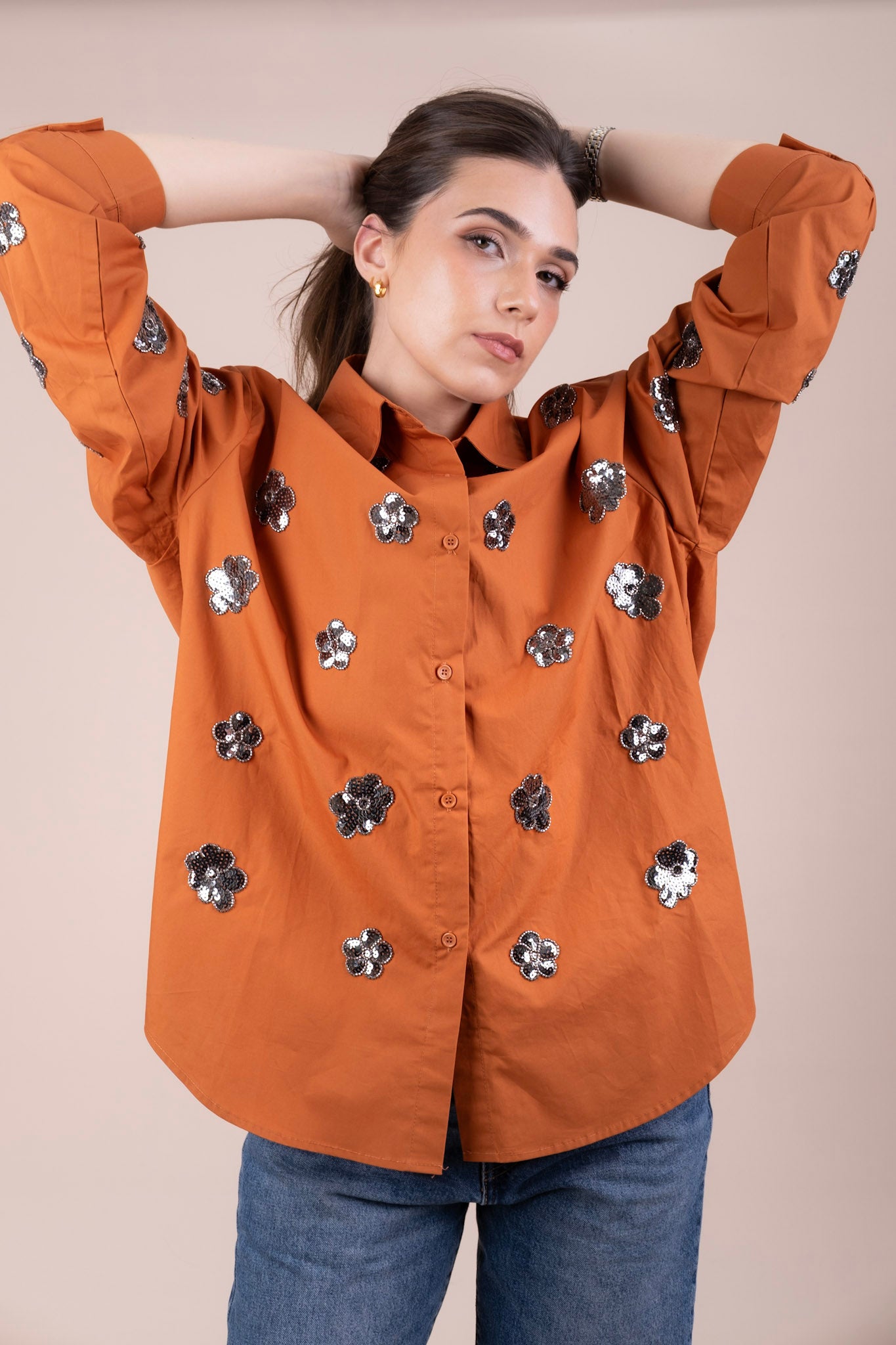 A woman wearing a burnt orange button-up shirt with silver sequined flower appliqués, paired with blue jeans, posing with her hands behind her head against a solid beige background.