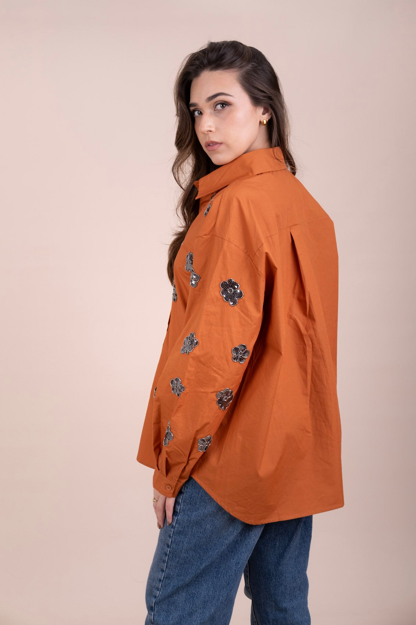 A woman wearing an oversized burnt orange button-up full sleeves shirt with silver sequined floral embellishments