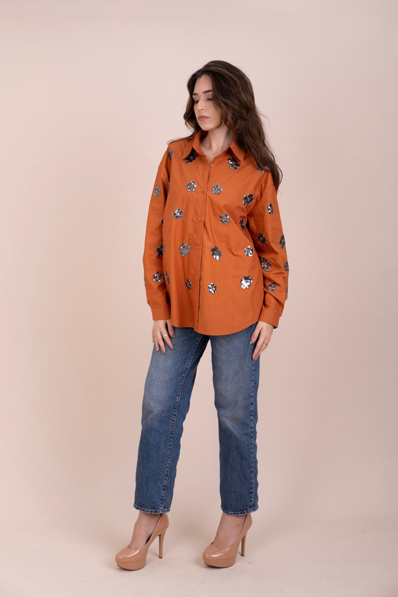 A woman modelling a burnt orange long-sleeve button-up shirt with silver floral embroidery, paired with blue jeans and beige high heels.