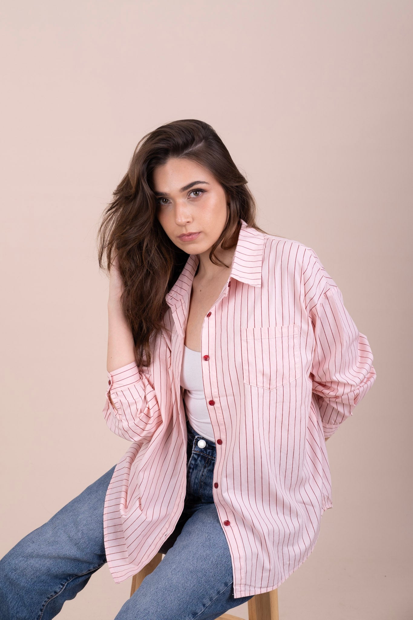 A woman wearing a pink and white vertically striped button-up shirt over a white tank top and blue jeans, seated against a solid beige studio background.