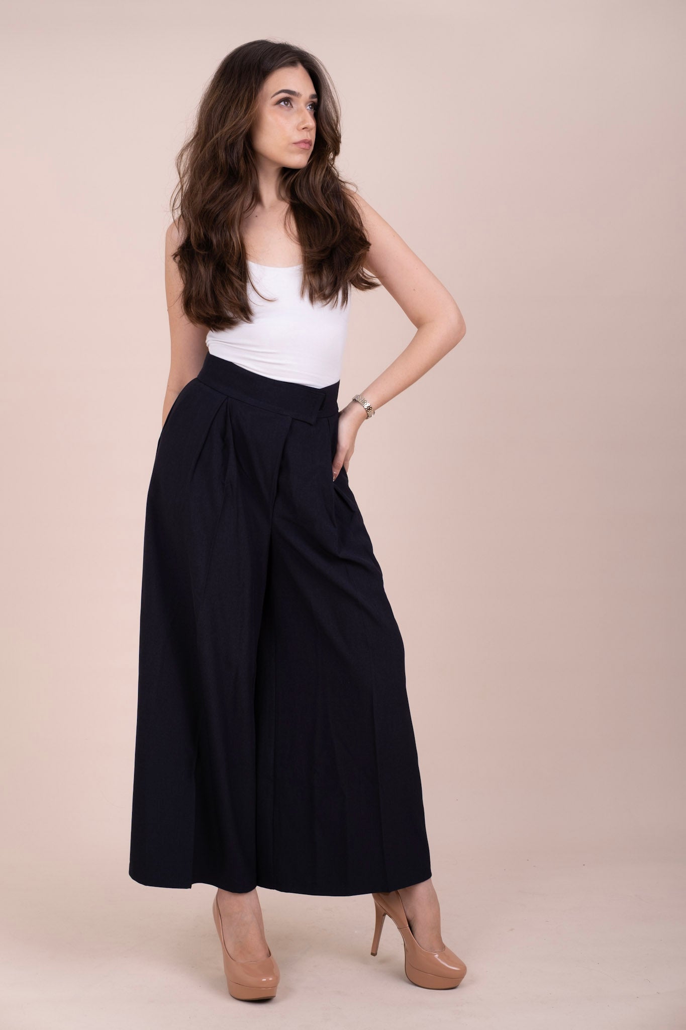 A woman modeling a pair of high-waisted, wide-leg navy blue pants with a white strapless top and nude high heels, set against a solid beige studio background.