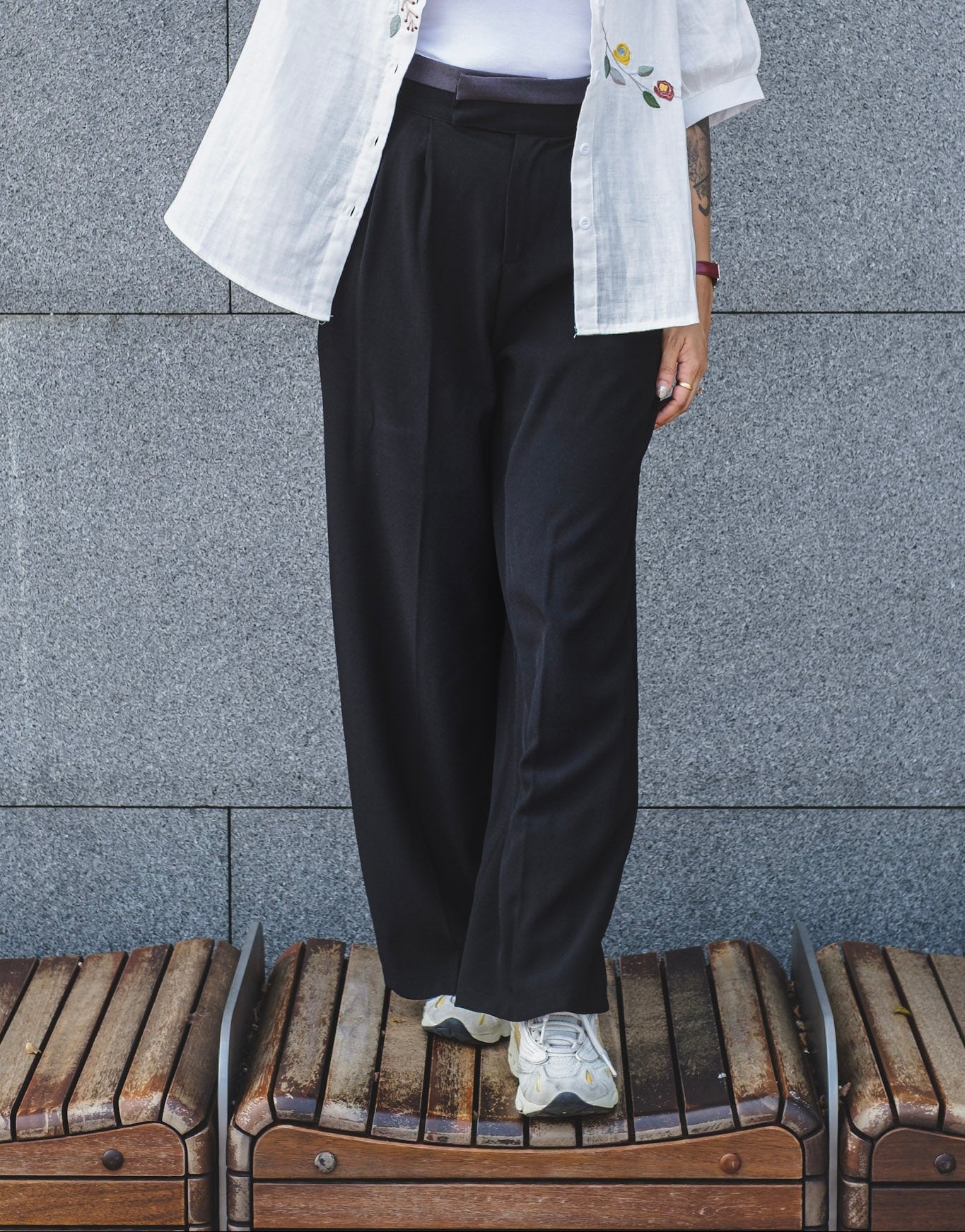 A person wearing a black wide-leg pants, and white sneakers, standing on wooden benches against a gray stone wall.