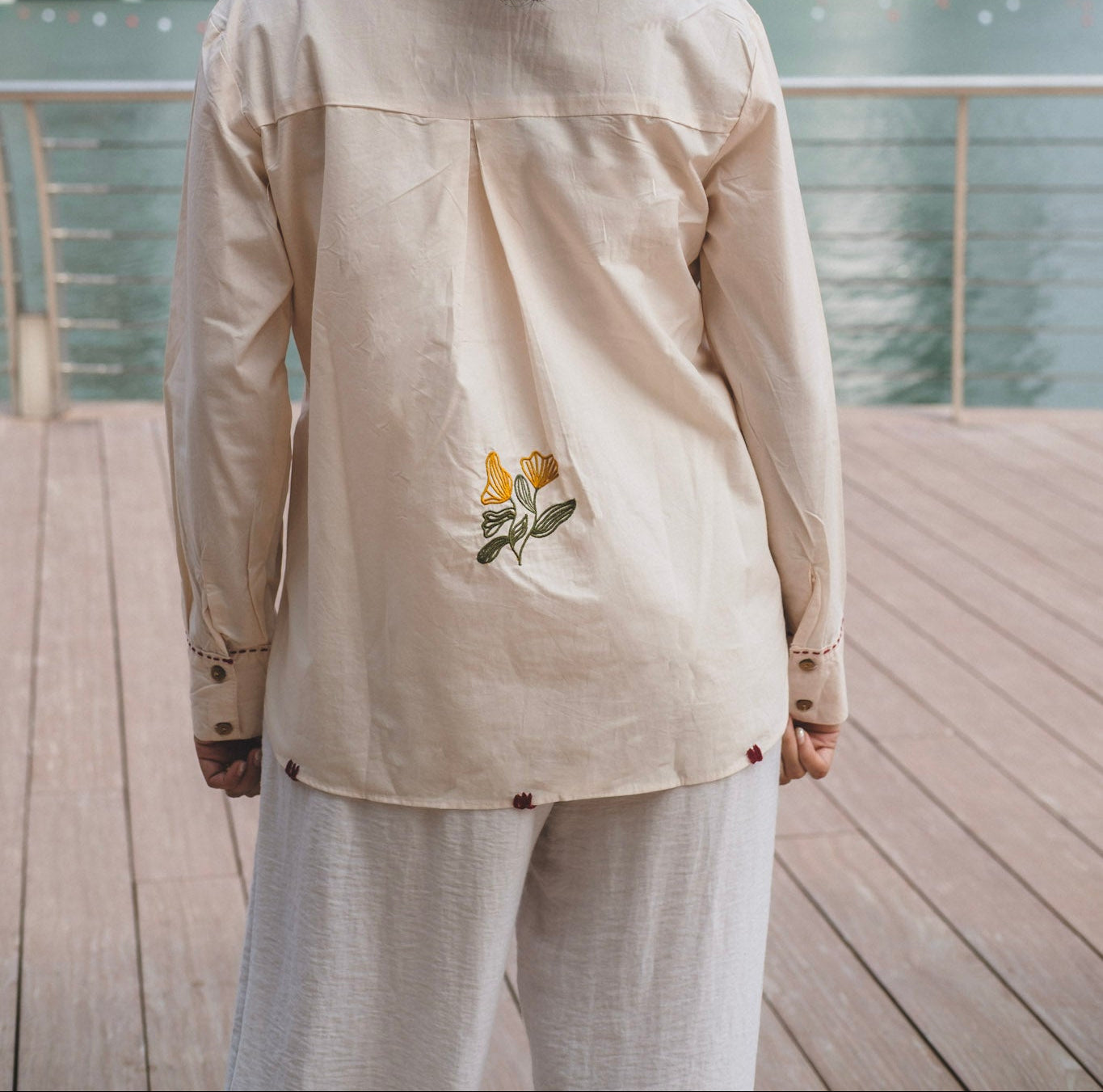 Woman viewed from behind wearing a light beige cotton button-up shirt with yellow and green floral embroidery on the back, standing on a wooden deck.