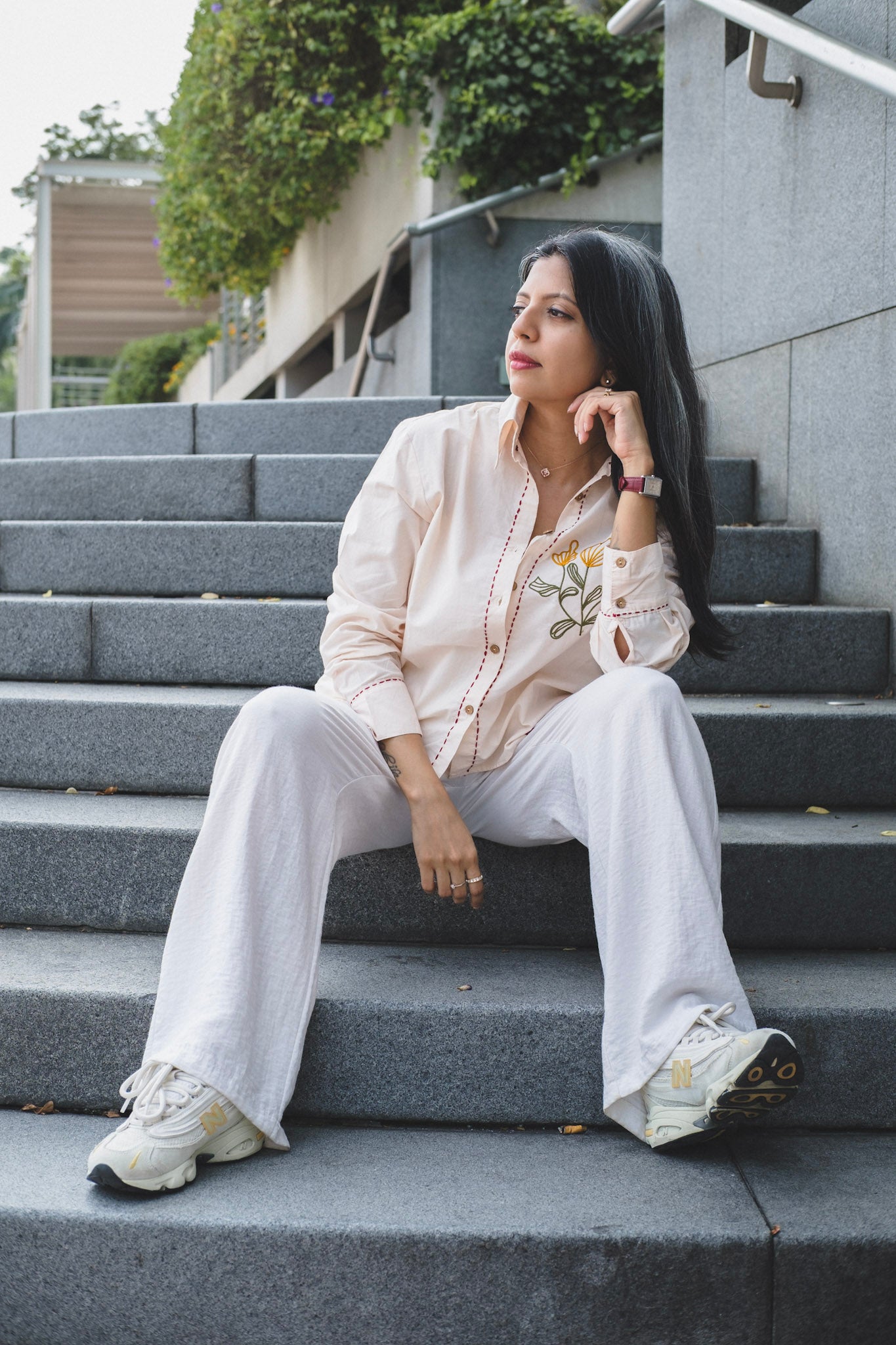 A woman sits on outdoor stone steps wearing a light beige embroidered button-up shirt with floral embroidery, matching wide-leg trousers, and white sneakers.